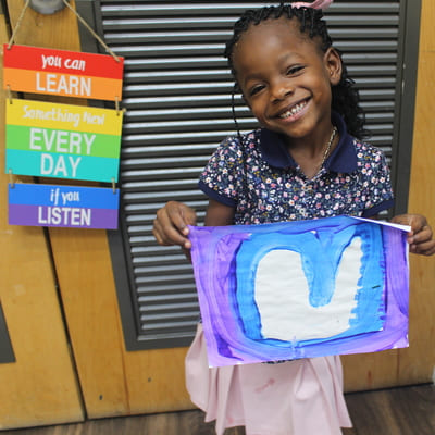 A little girl holding an art blue and purple drawing while standing and smiling next to a colorful sign