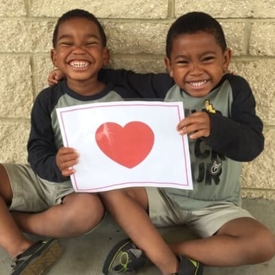 Young boy twins hugging each other while sitting cross legged on the floor and holding a white piece of paper with a red heart in the middle of it